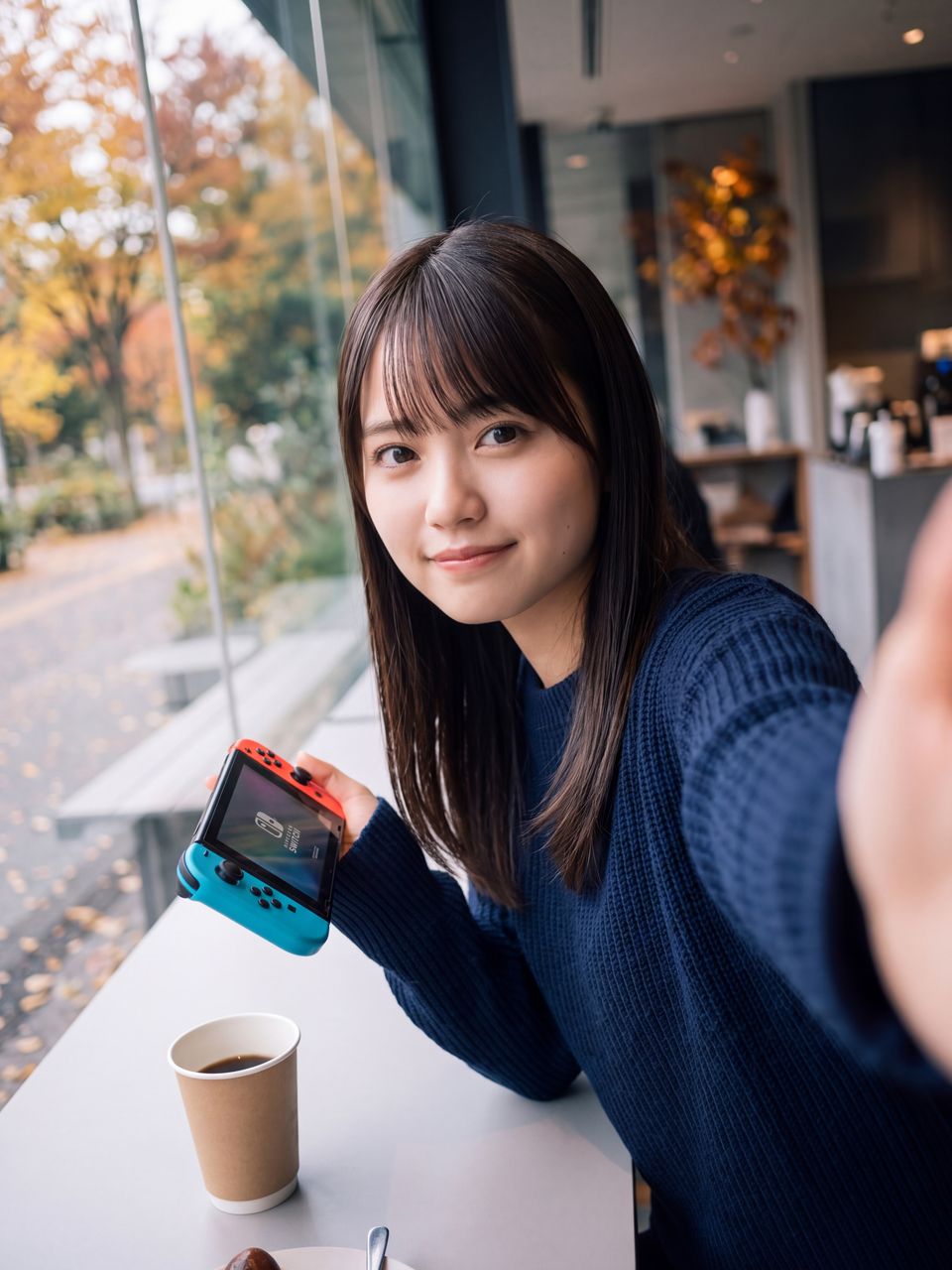 カフェの窓際席で携帯ゲーム機を手に控えめな微笑みを浮かべる蒼井瑠璃のセルフィーショット