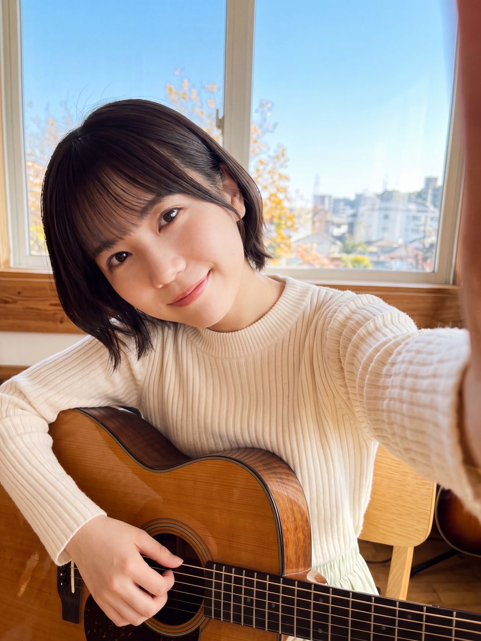 秋晴れのスタジオの窓辺でアコースティックギターを膝に抱えて満面の笑みで微笑む天音かなでの自撮り写真