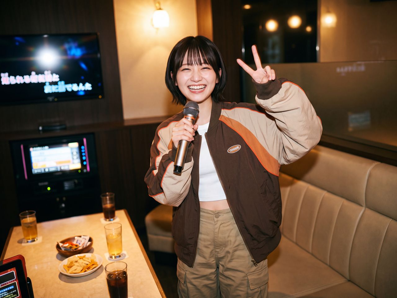 カラオケルームでワイヤレスマイクを手に持ち、秋のスポーティーカジュアルなジャケット姿で元気いっぱいにピースサインをして満面の笑みを浮かべる神楽坂舞
